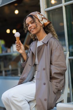happy african american woman in headscarf holding ice cream cone and sitting on chair in prague