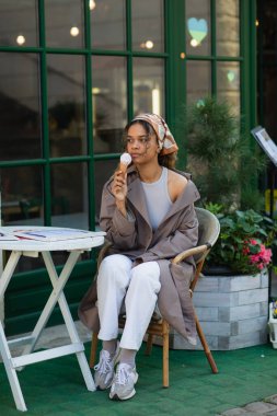 young african american woman in headscarf and stylish trench coat holding ice cream cone and sitting in cafe terrace 