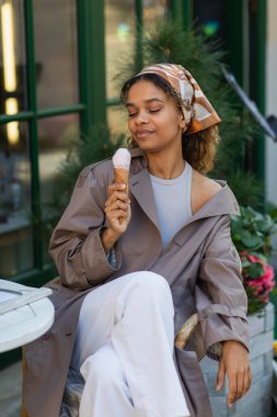 pleased african american woman in headscarf and stylish trench coat holding ice cream cone and sitting in cafe terrace 