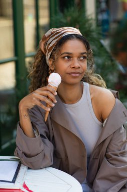 tattooed african american woman in headscarf holding ice cream cone and sitting in cafe terrace 