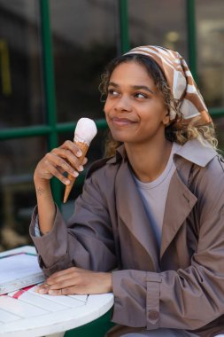 smiling african american woman in stylish trench coat holding ice cream cone and sitting in cafe terrace 