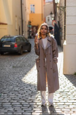 full length of dreamy african american woman in trench coat and headscarf holding ice cream cone on street in prague 