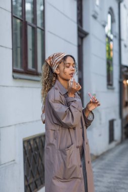 curly african american woman in stylish trench coat and headscarf applying lip gloss on street in prague 