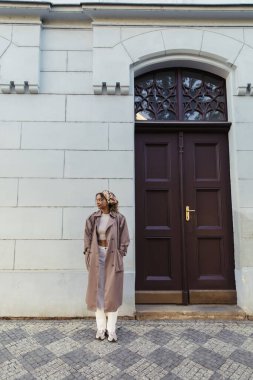 full length of african american woman in trendy outfit and headscarf standing with hands in pockets near building in prague