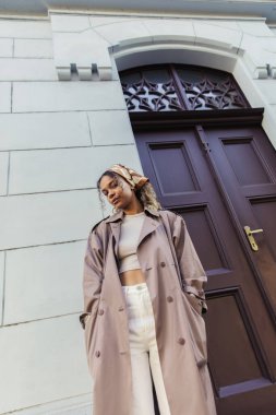 low angle view of african american woman in trendy outfit and headscarf standing with hands in pockets near building in prague