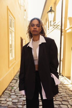 stylish african american woman in trendy oversize suit strolling with hand in pocket on street in prague