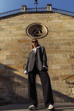 low angle view of trendy african american woman in sunglasses and oversize suit standing near ancient wall on street in prague