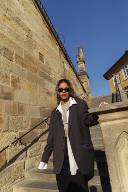 stylish african american woman in sunglasses and oversize suit walking on stairs on street in prague 