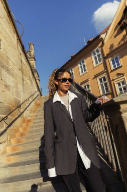 stylish african american woman in sunglasses and oversize suit on stairs of ancient street in prague 