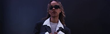 stylish african american woman in sunglasses and oversize suit near dark blue wall, banner