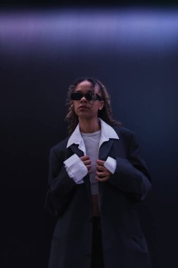 curly african american woman in sunglasses adjusting oversize blazer near dark blue wall