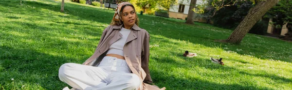 young african american woman in headscarf and stylish trench coat resting on green grass in park, banner