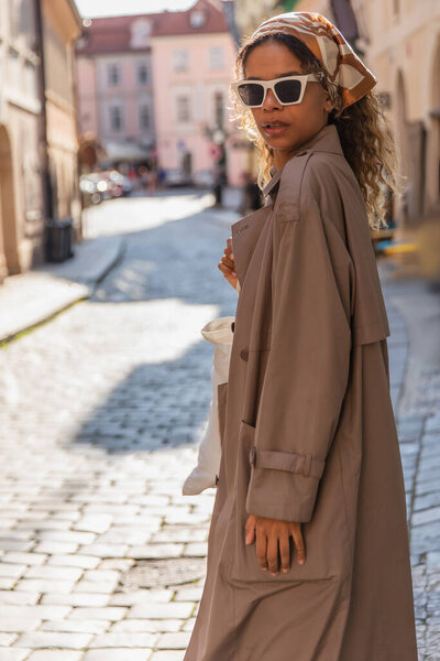 stylish african american woman in headscarf and sunglasses walking in trench coat on street in prague 