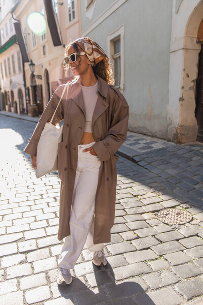 full length of happy african american woman in trendy accessories posing with hand in pocket on street in prague