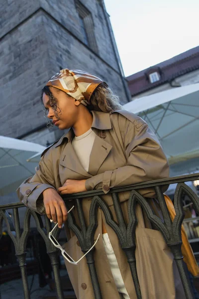 young african american woman in stylish outfit and headscarf holding sunglasses on street in prague 