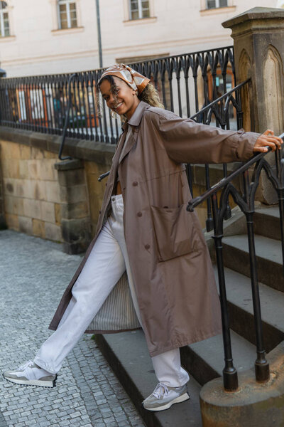 full length of happy african american woman in trench coat standing on stairs 