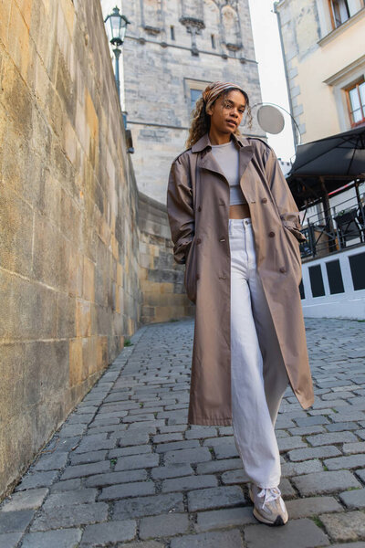 Полная длина стильный африканский американец женщина в модном плаще ходить с руками в карманах возле древней стены в Праге