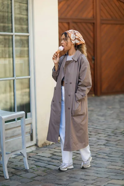 curly african american woman in headscarf and stylish trench coat eating ice cream cone on street in prague 