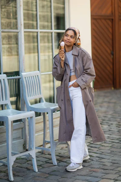full length of african american woman in headscarf and trench coat holding ice cream cone and walking on street in prague 