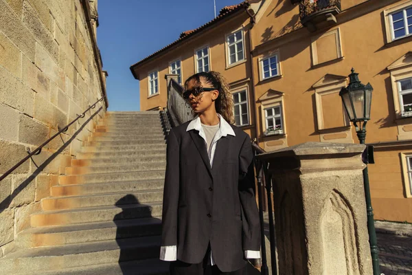 stylish african american woman in sunglasses and oversize suit standing near stairs on street in prague 