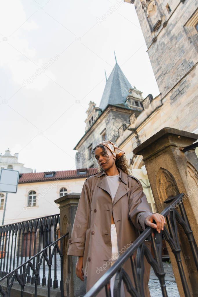 low angle view of stylish african american woman in trench coat near old town hall tower in prague
