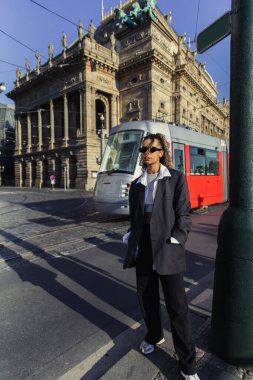 stylish african american woman in oversized blazer and sunglasses standing with hands in pockets near national theatre in prague clipart