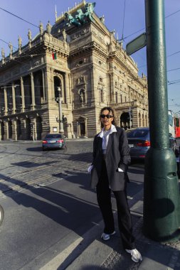stylish african american woman in oversized blazer and sunglasses standing with hands in pockets near national theatre in prague