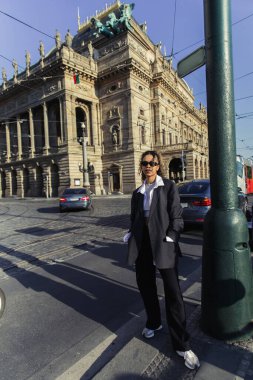 stylish african american woman in oversized blazer and sunglasses standing with hands in pockets near national theatre in prague clipart