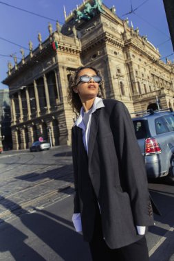stylish african american woman in oversized blazer and sunglasses standing near national theatre in prague