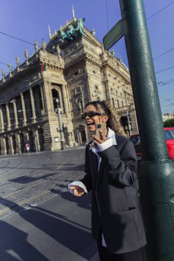 happy african american woman in oversized blazer and sunglasses holding smartphone near national theatre in prague