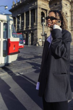 happy african american woman in oversized blazer and sunglasses talking on smartphone in prague