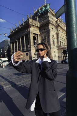 curly african american woman in oversized blazer and sunglasses taking selfie near national theatre in prague