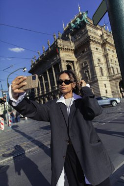 young african american woman in oversized blazer and sunglasses taking selfie near national theatre in prague clipart