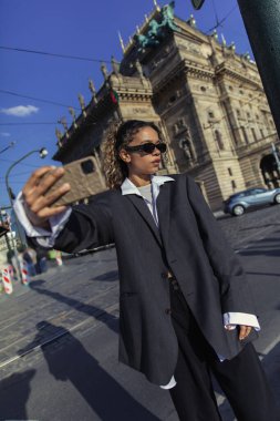 stylish african american woman in oversized blazer and sunglasses taking selfie near national theatre in prague clipart