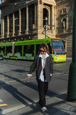 young african american woman in stylish blazer and sunglasses walking with hand in pocket near tram and building in prague