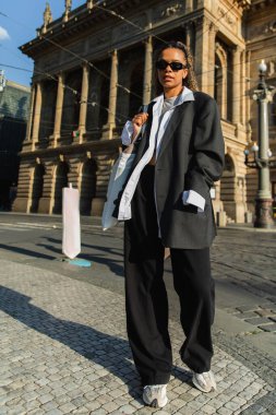full length of young african american woman in sunglasses walking near national theatre in prague clipart