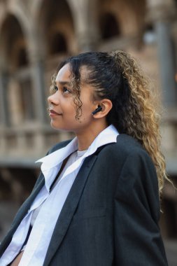 side view of dreamy african american woman in oversize suit listening music in wireless earphone on street of prague 