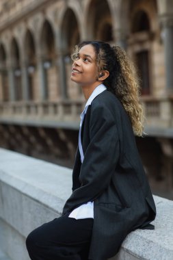 pleased african american woman in oversize suit listening music in wireless earphone on urban street of prague 