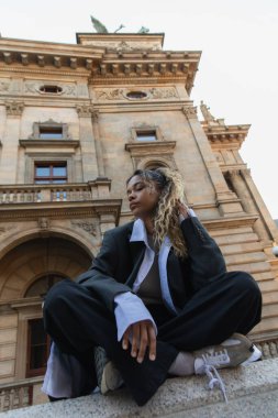 low angle view of african american woman in oversize suit sitting with crossed legs near national theatre in prague  clipart
