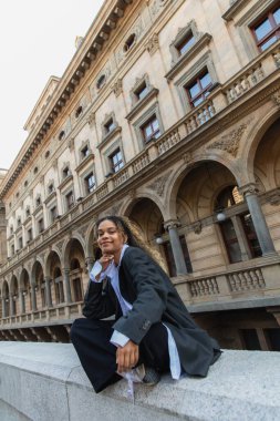pleased african american woman in oversize suit listening music in wireless earphone near building in prague 