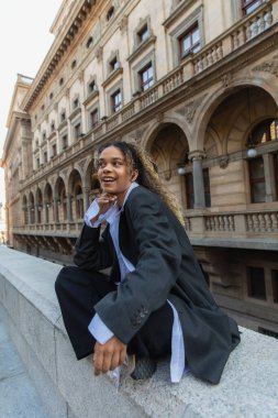 amazed african american woman in oversize suit listening music in wireless earphone near building in prague 