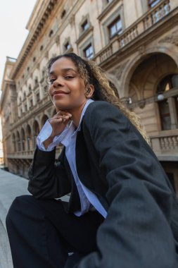 smiling african american woman in oversize suit listening music in wireless earphone near building in prague 