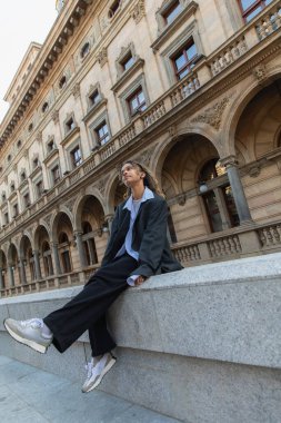 african american woman in oversize suit listening music in wireless earphone wile sitting near building in prague 
