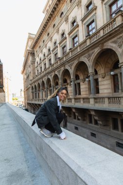 positive african american woman in oversize suit listening music in wireless earphone while sitting near building in prague 