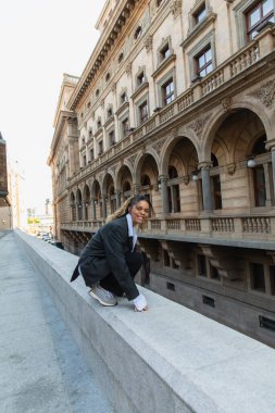 positive african american woman in oversize suit listening music in wireless earphone while sitting near building in prague  clipart