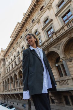 african american woman in blazer listening music in wireless earphone on urban street near building of national theater in prague