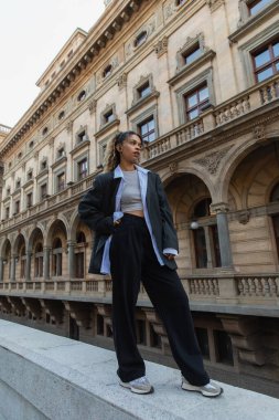 african american woman in wireless earphone posing on urban street near building of national theater in prague