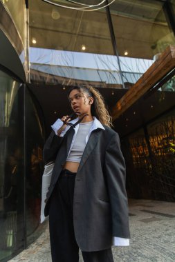 trendy african american woman in wireless earphone holding sunglasses on urban street in prague 