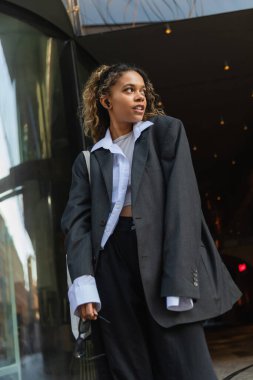 african american woman in oversize blazer holding sunglasses while listening music on urban street in prague 