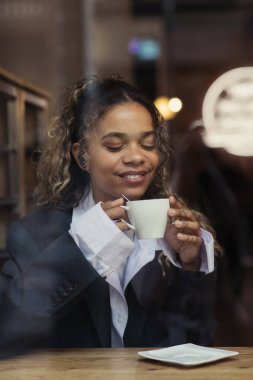 pleased african american woman in wireless earphone listening music and holding cup of coffee in cafe